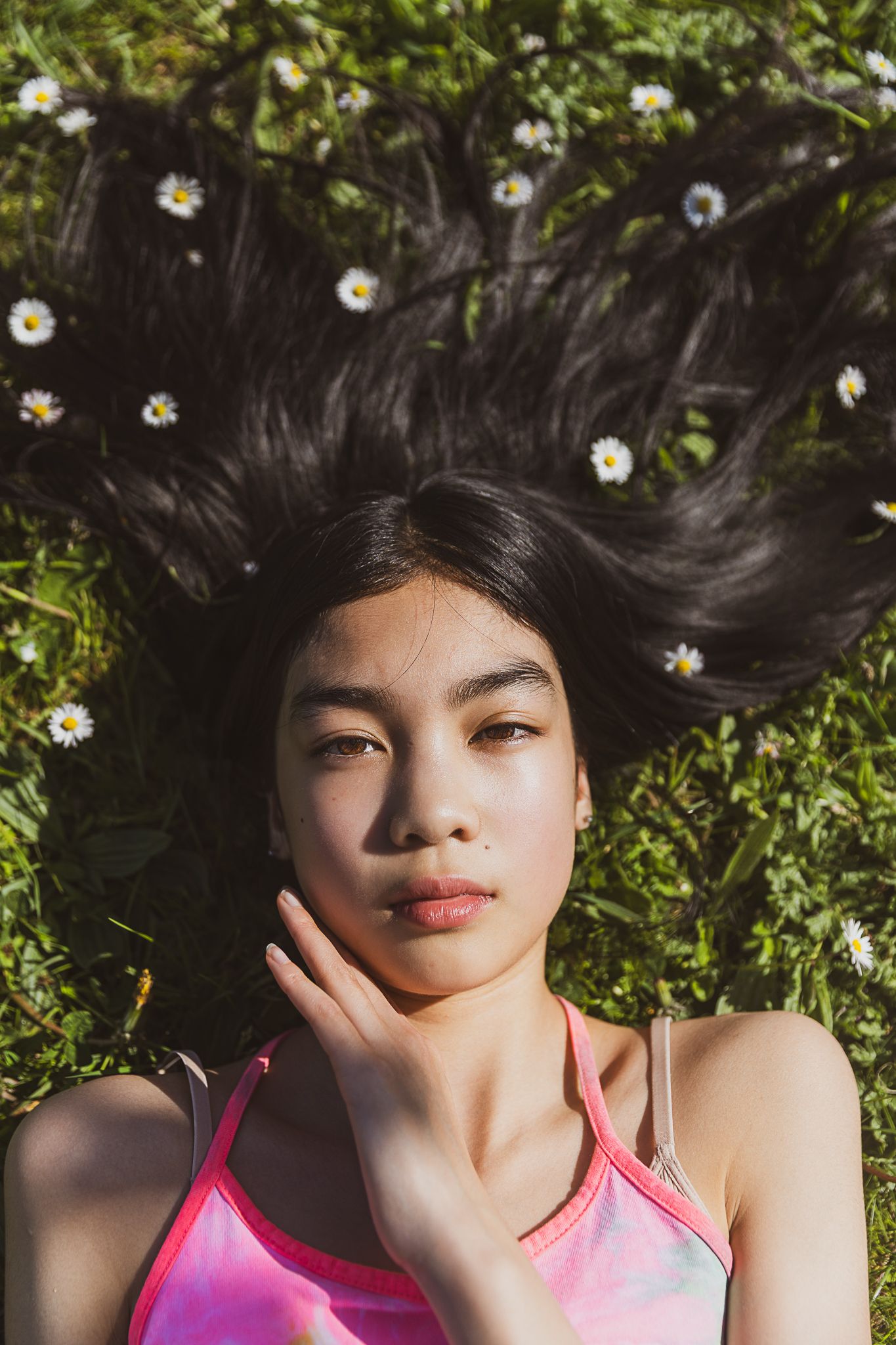 A person laying in grass surrounded by small white daisies, wearing a pink tank top and looking up at the sky. Kinderkledingmerk fotografie