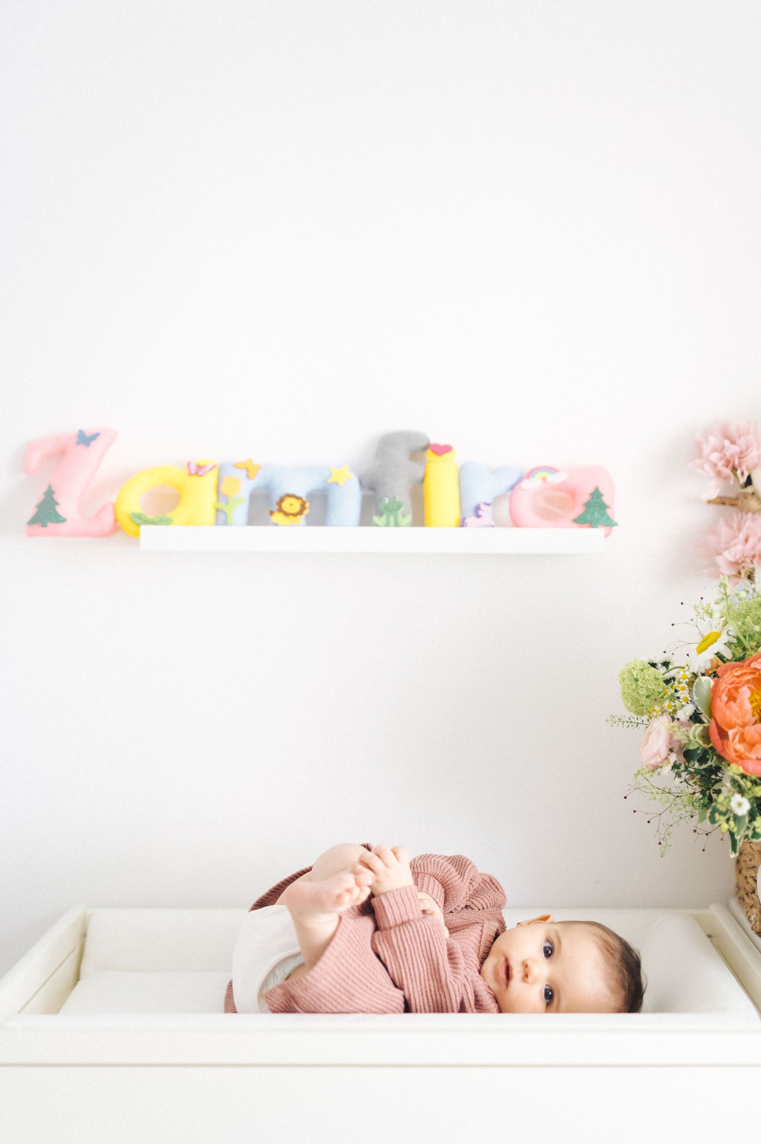 Baby in pink clothing lies on white changing table with colorful books on shelf and floral arrangement nearby.