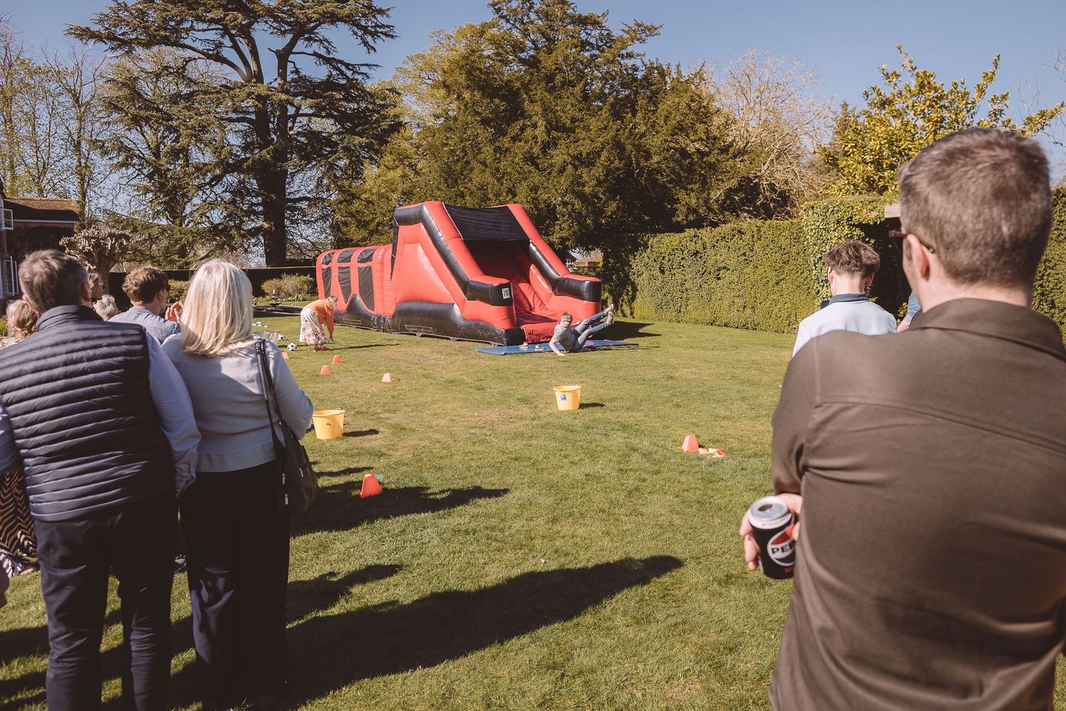 People gather on a lawn watching a large red inflatable bounce house during an outdoor event on a sunny day.
