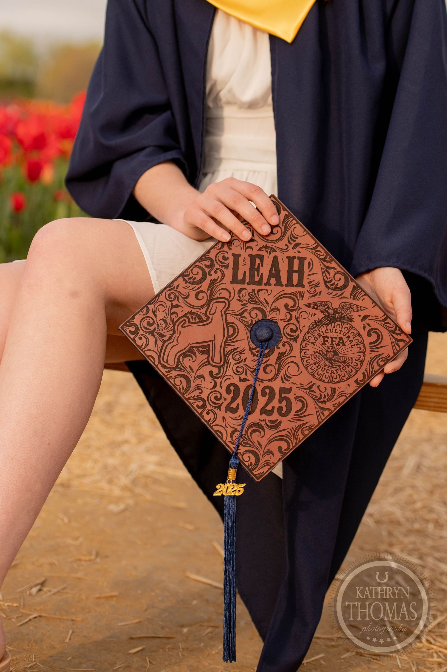 Personalized brown leather graduation cap with ornate design featuring 'LEAH 2025' decorated with floral patterns and tassel.