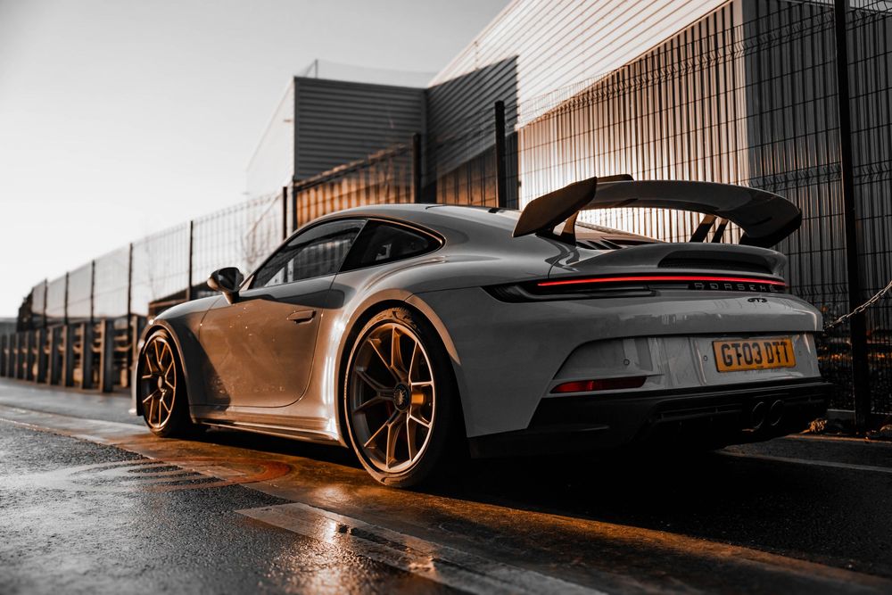 Gray Porsche 911 GT3 rear view showcasing large rear wing and bronze wheels in wet conditions.