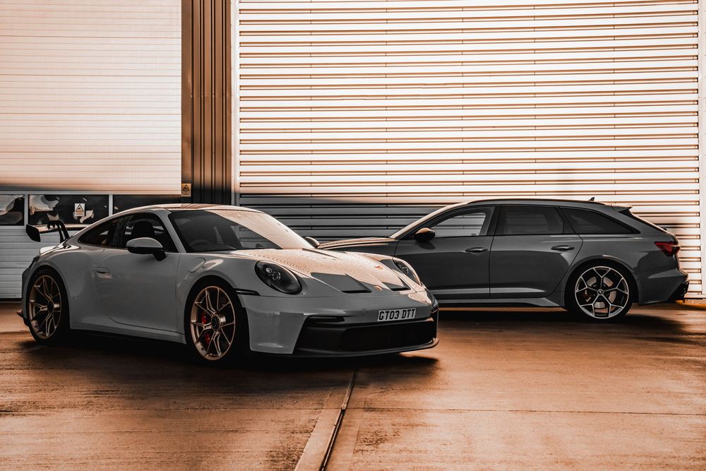 A silver Porsche 911 GT3 and dark Audi RS wagon parked side by side in front of metal garage doors.