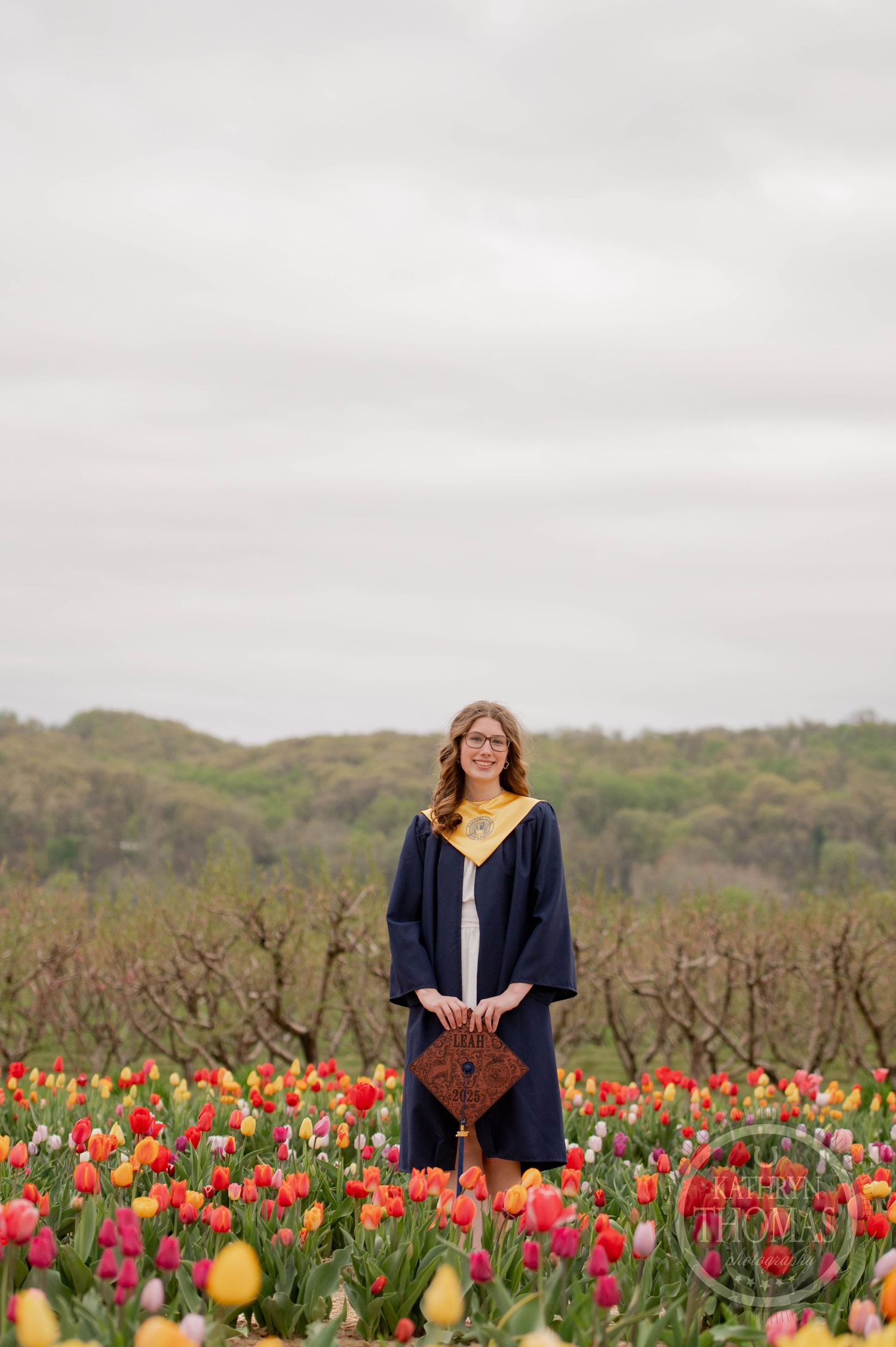 Woman in dark cardigan stands in colorful tulip field on overcast day with hills in background.