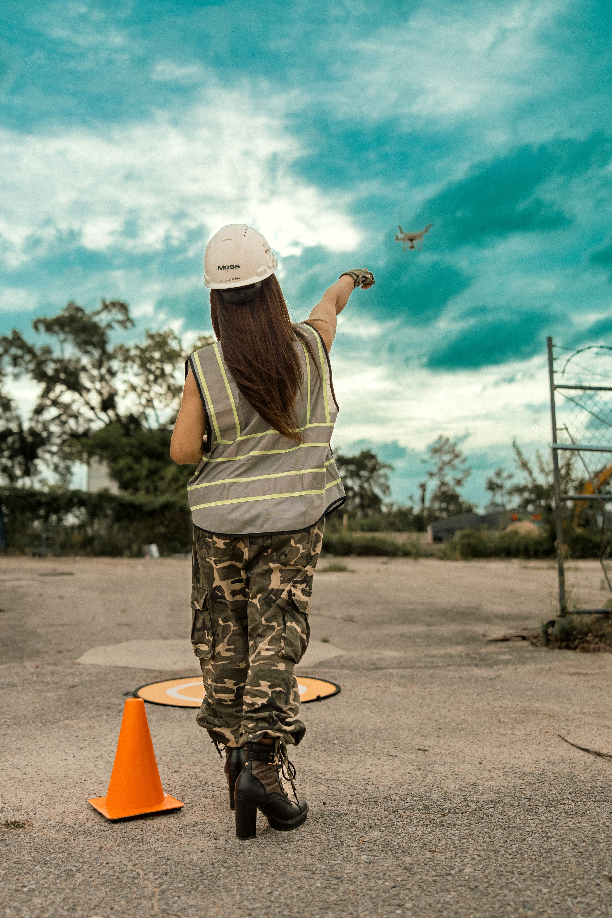 Drone operator in safety vest and camo pants controls aircraft against turquoise sky with orange traffic cone.