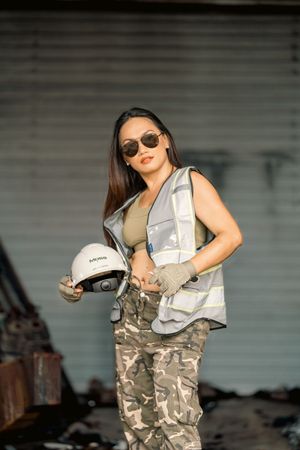 Industrial worker in safety equipment holds hard hat while posing near construction site.