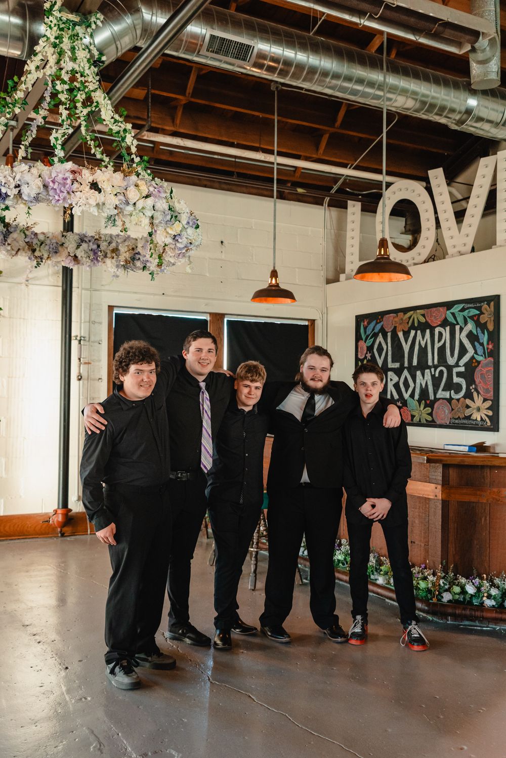A group in formal black attire poses together in an industrial-style venue with exposed ductwork and decorative lighting.