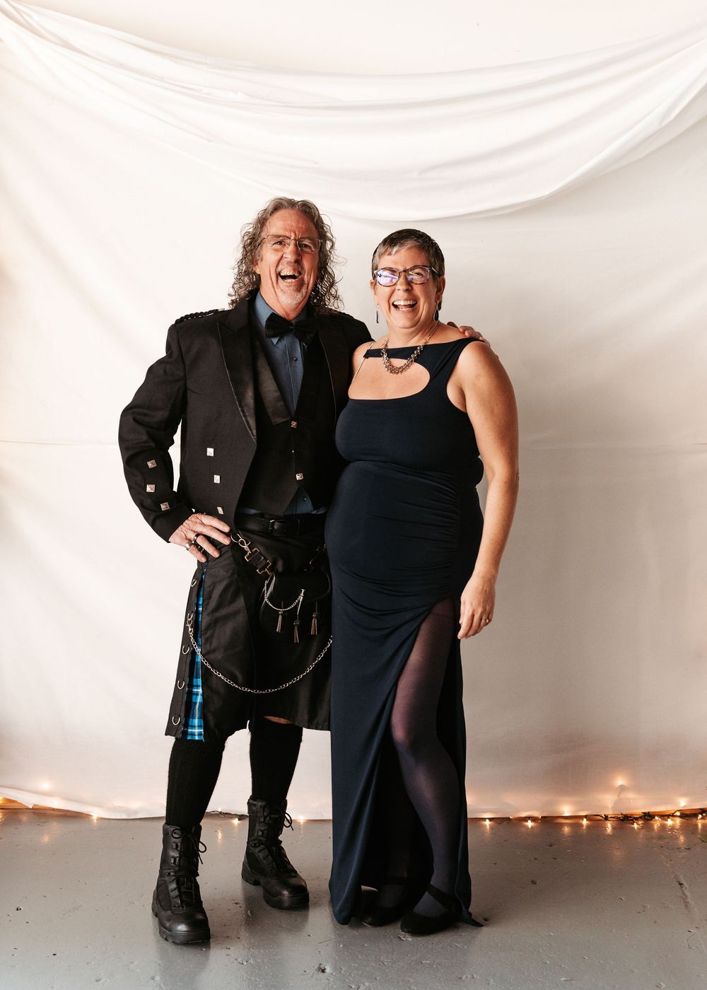 A couple poses together against a white backdrop with string lights, one wearing a black kilt and the other in a formal black dress.