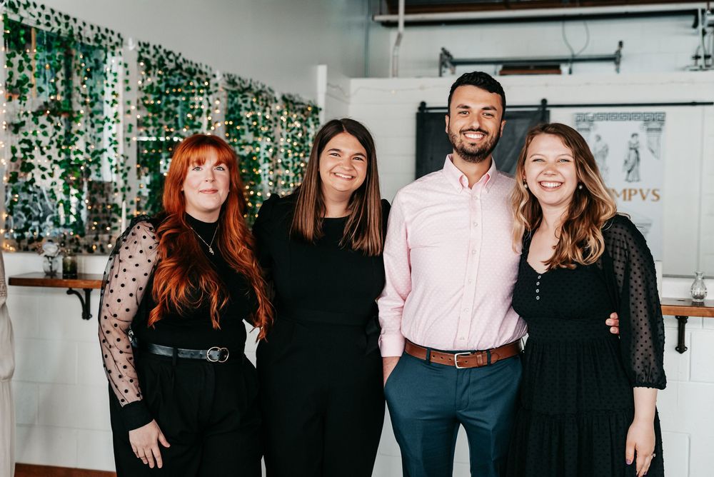 A group of four people stand together smiling in an office space with a green floral wall backdrop.