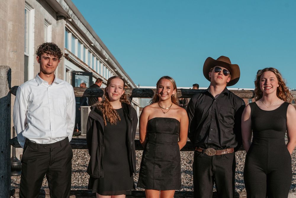 Group of people in formal black and white attire posing together outdoors against a building on a sunny day.
