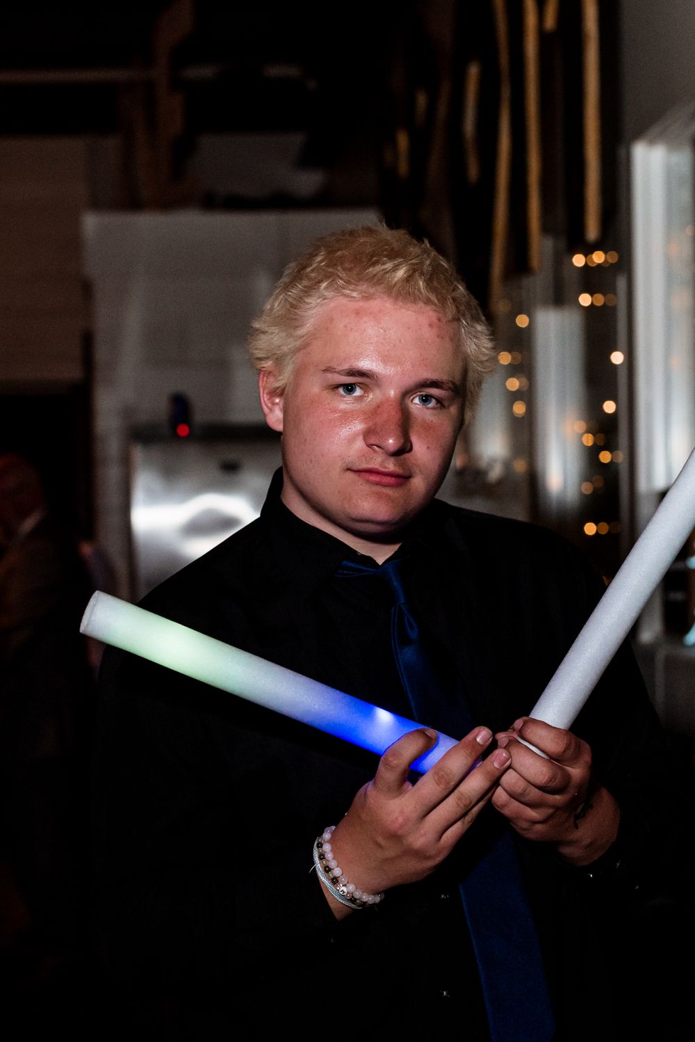 Person in a dark suit holds illuminated blue and white light-up sticks in a dimly lit room with twinkle lights behind.