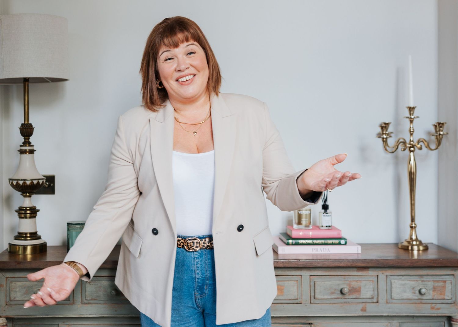 A person in a white blazer and blue dress stands gesturing in an elegant room with decorative candelabras.