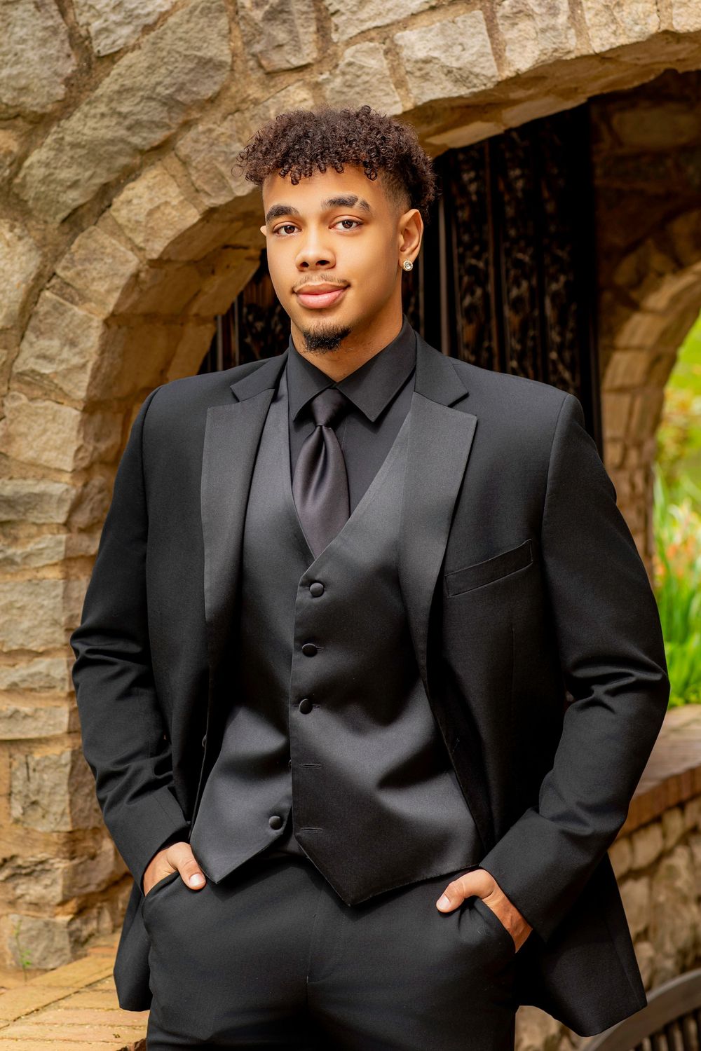 Young man in formal black three piece suit and tie poses confidently in stone archway.