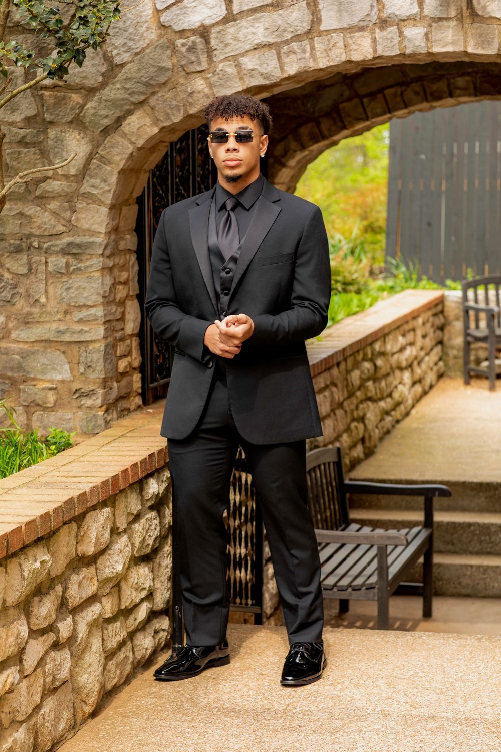 Stylish figure in all-black formal attire poses confidently under stone archway at outdoor venue.