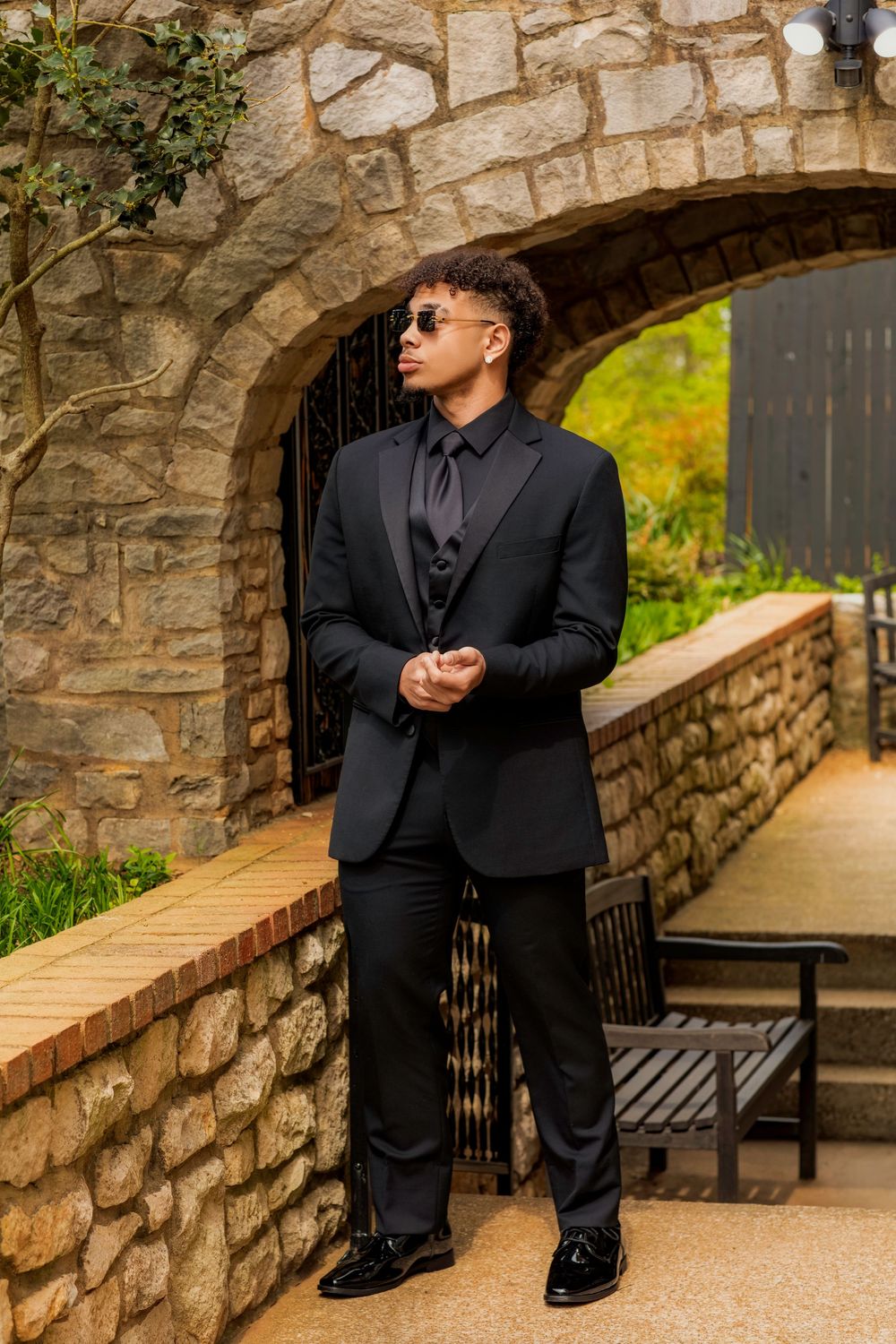 A stylish all-black formal outfit is showcased against a stone archway and garden path backdrop.