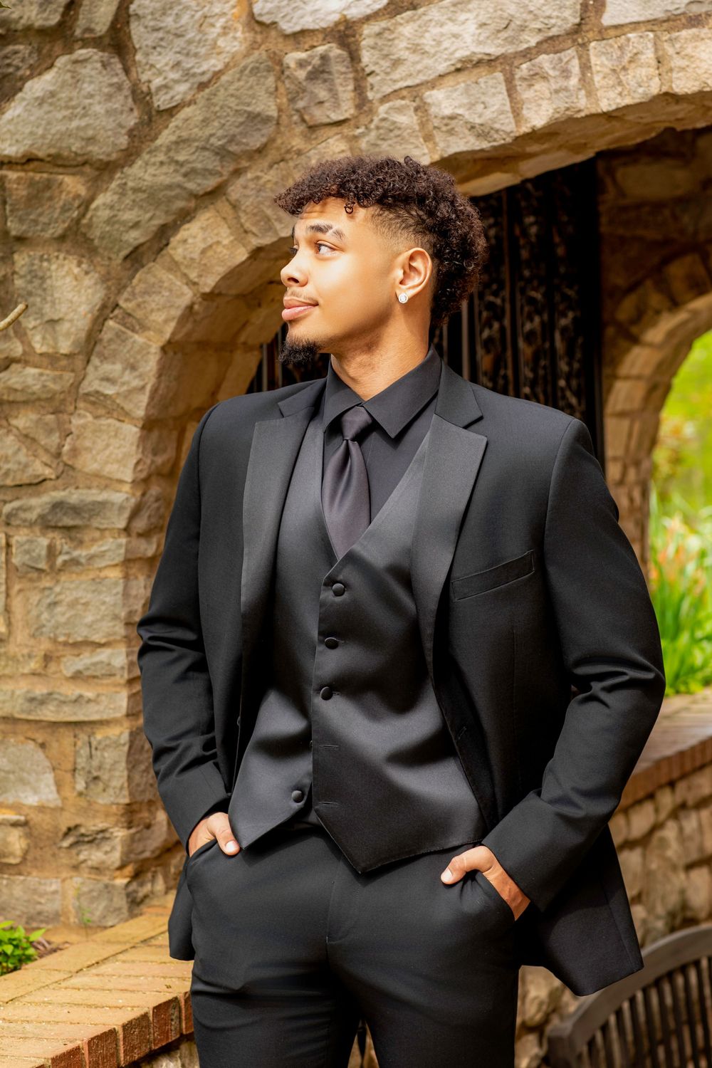 Young man in elegant black three-piece suit and tie poses against stone archway outdoors during formal event.