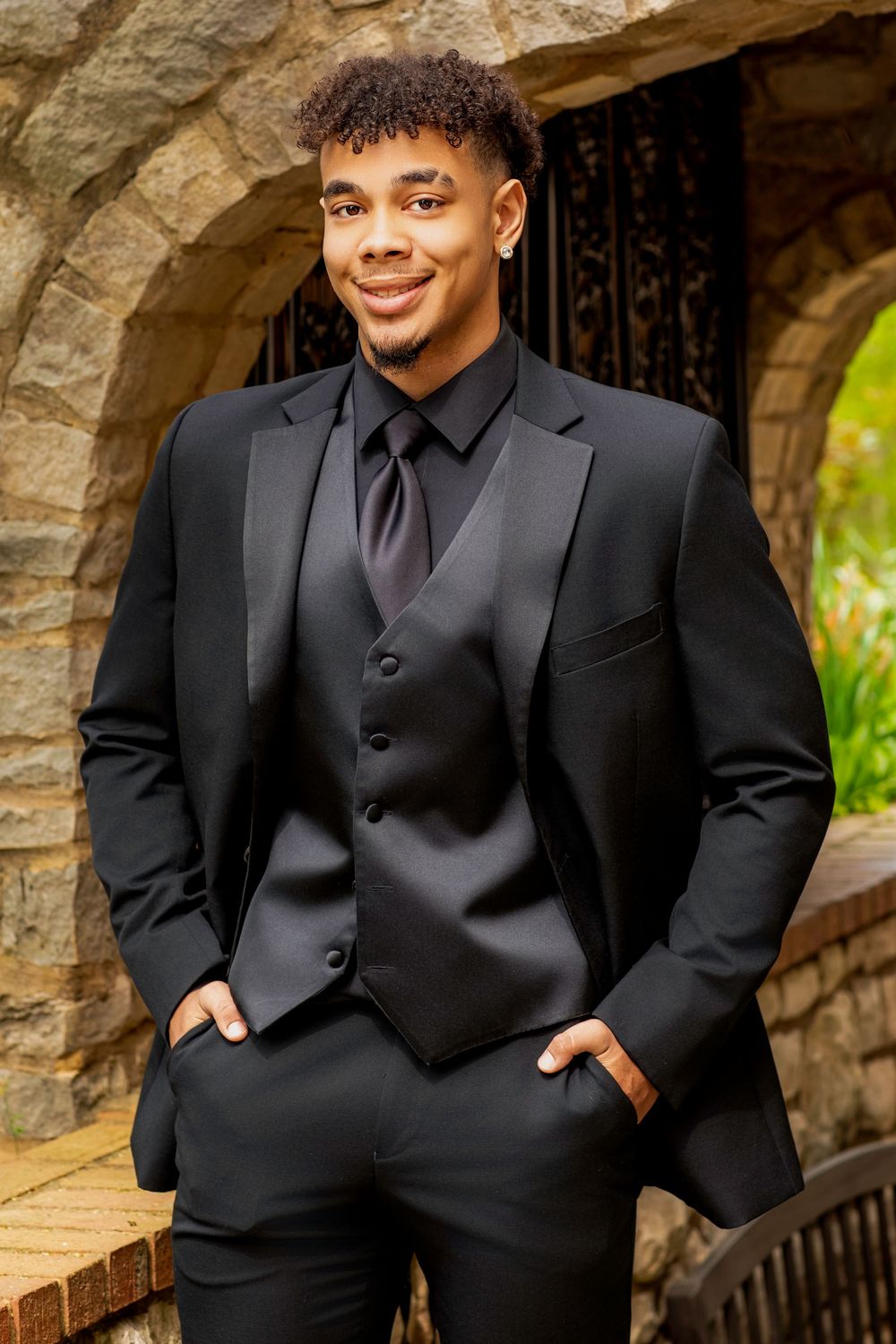 Well-dressed person in all-black formal attire including suit jacket, vest and tie poses confidently in front of stone archway.