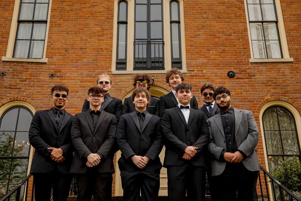 Group in formal black suits and bow ties standing in front of brick building with arched windows.