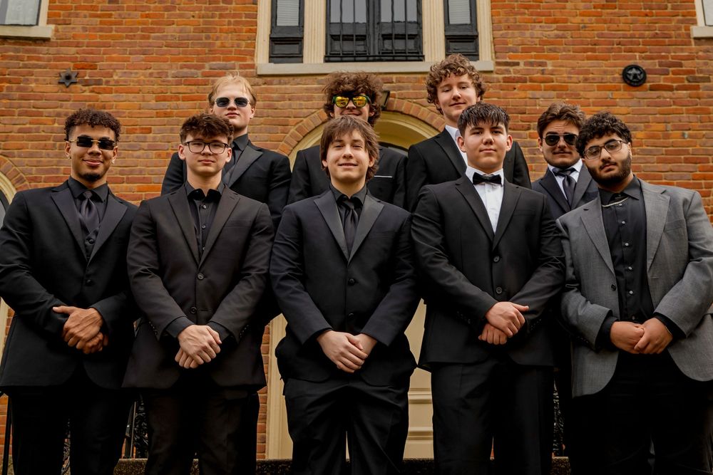 A group of groomsmen in black suits and tuxedos pose together in front of a brick building with an arched doorway.