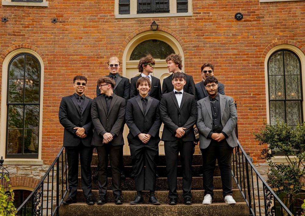A group in formal black suits and sunglasses stand together on brick building steps in matching attire.