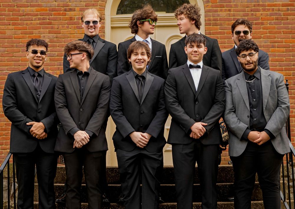 Group of formally dressed men in black and gray suits posing for photo in front of brick building.