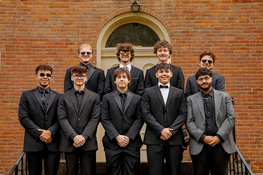 A group of men wearing formal dark suits and sunglasses pose together on steps of a brick building.