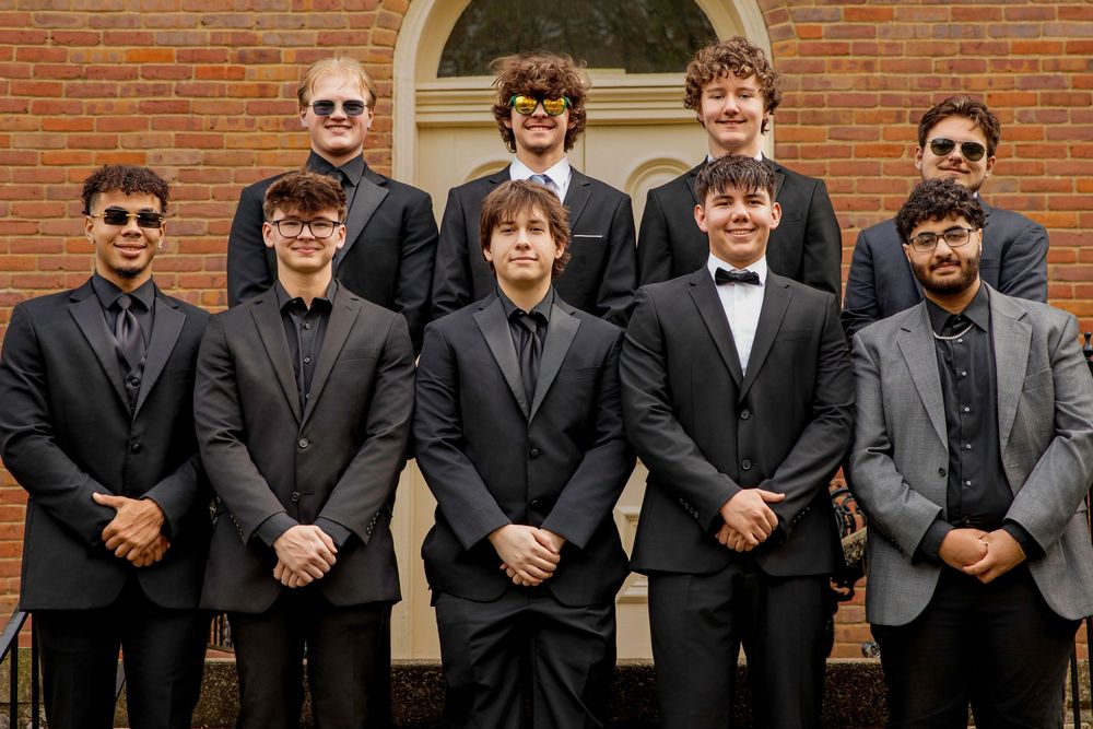 Group of men wearing black suits and sunglasses stand in a professional formation in front of brick building.
