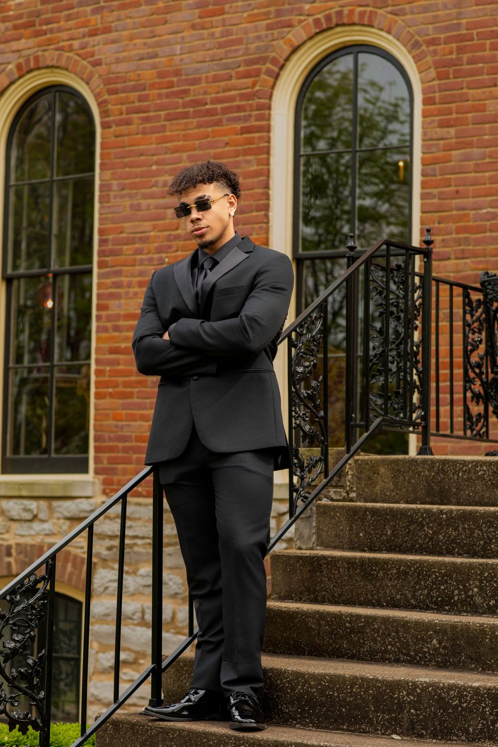 A person in a black suit and sunglasses stands confidently on stone steps in front of a brick building with arched windows.
