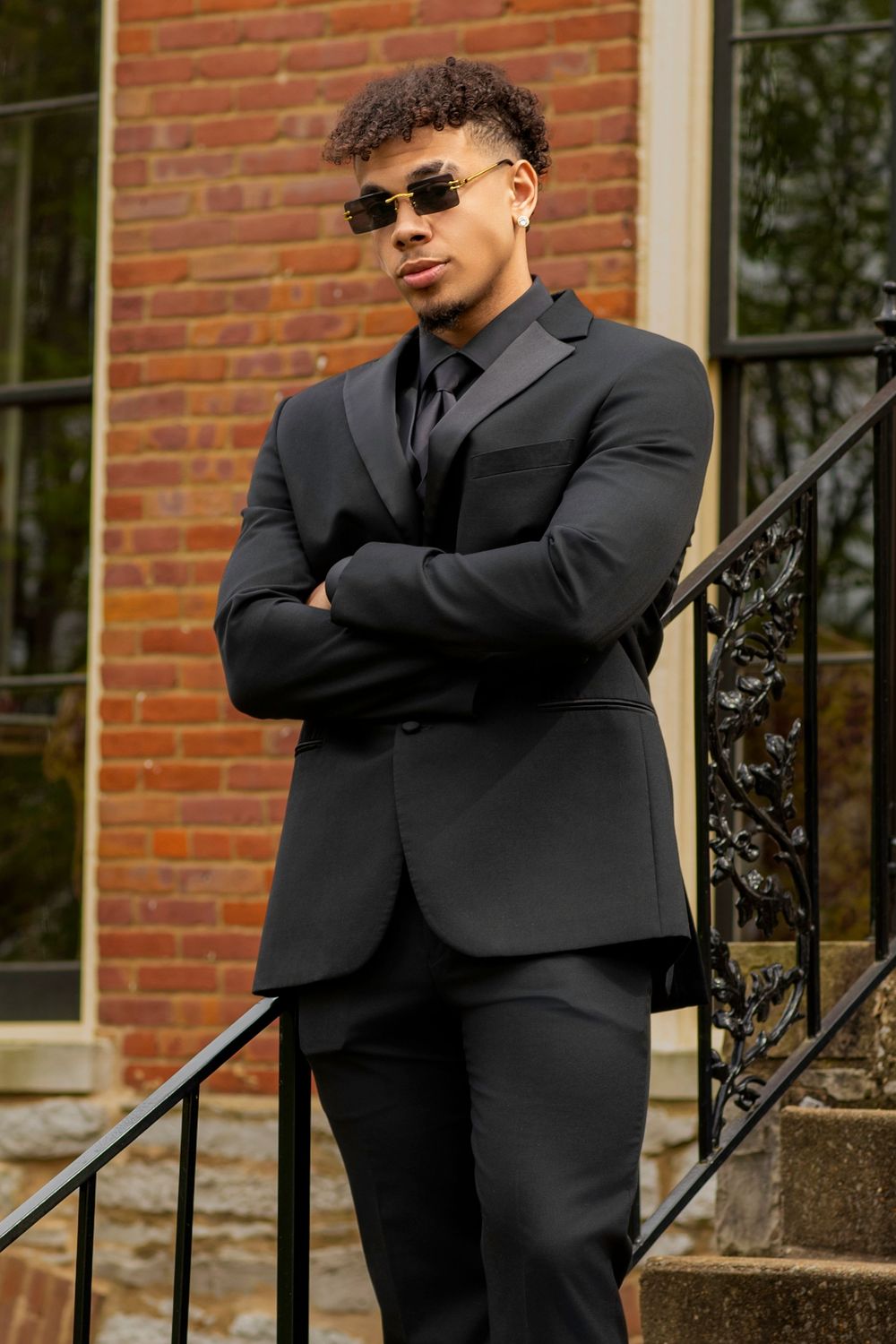 Sharp black suit with sunglasses standing confidently on brick building steps with ornate railing.