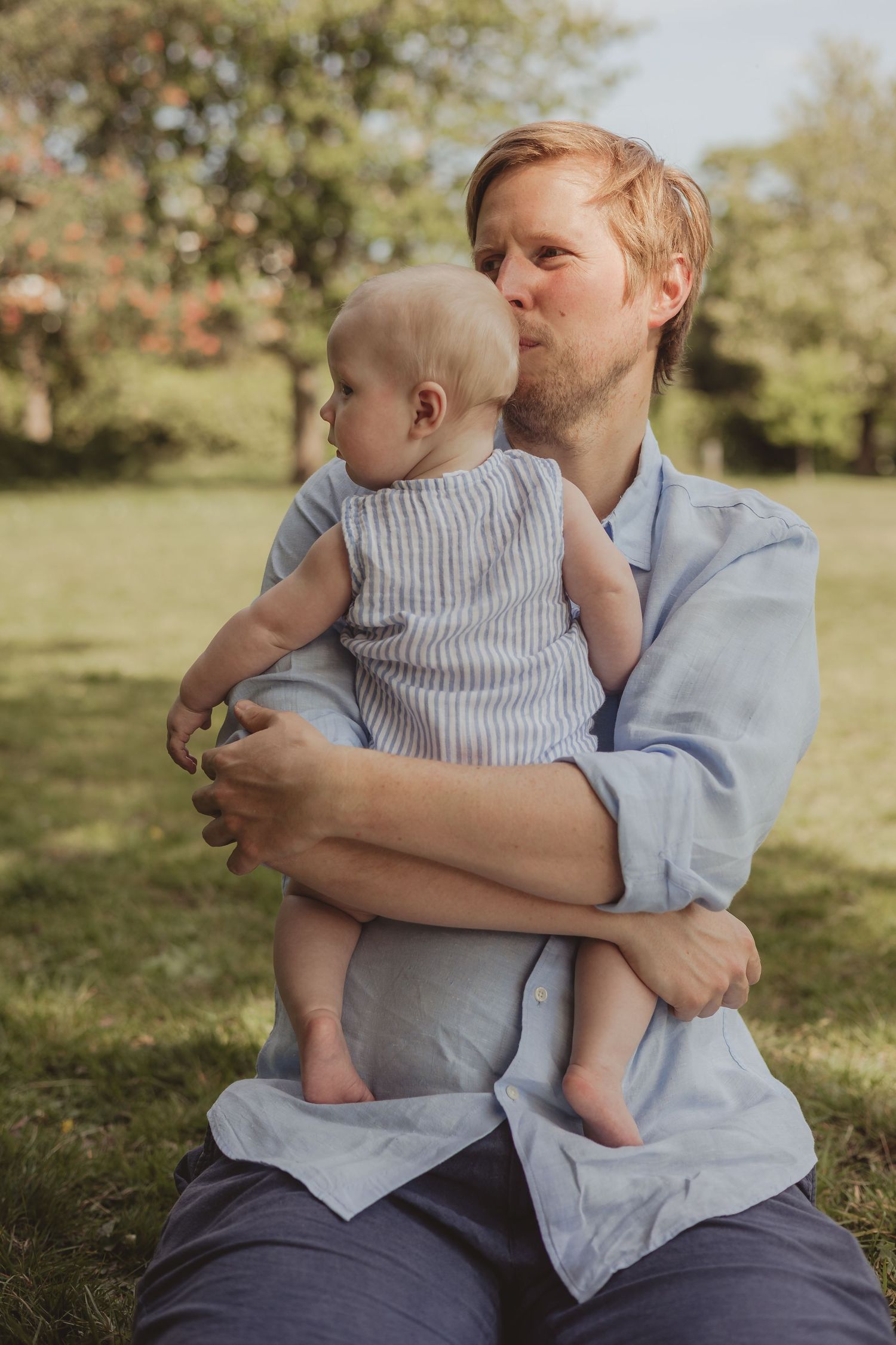 a father (dad) holds his baby to his chest on a sunny day in an outdoor park and kisses the baby's head