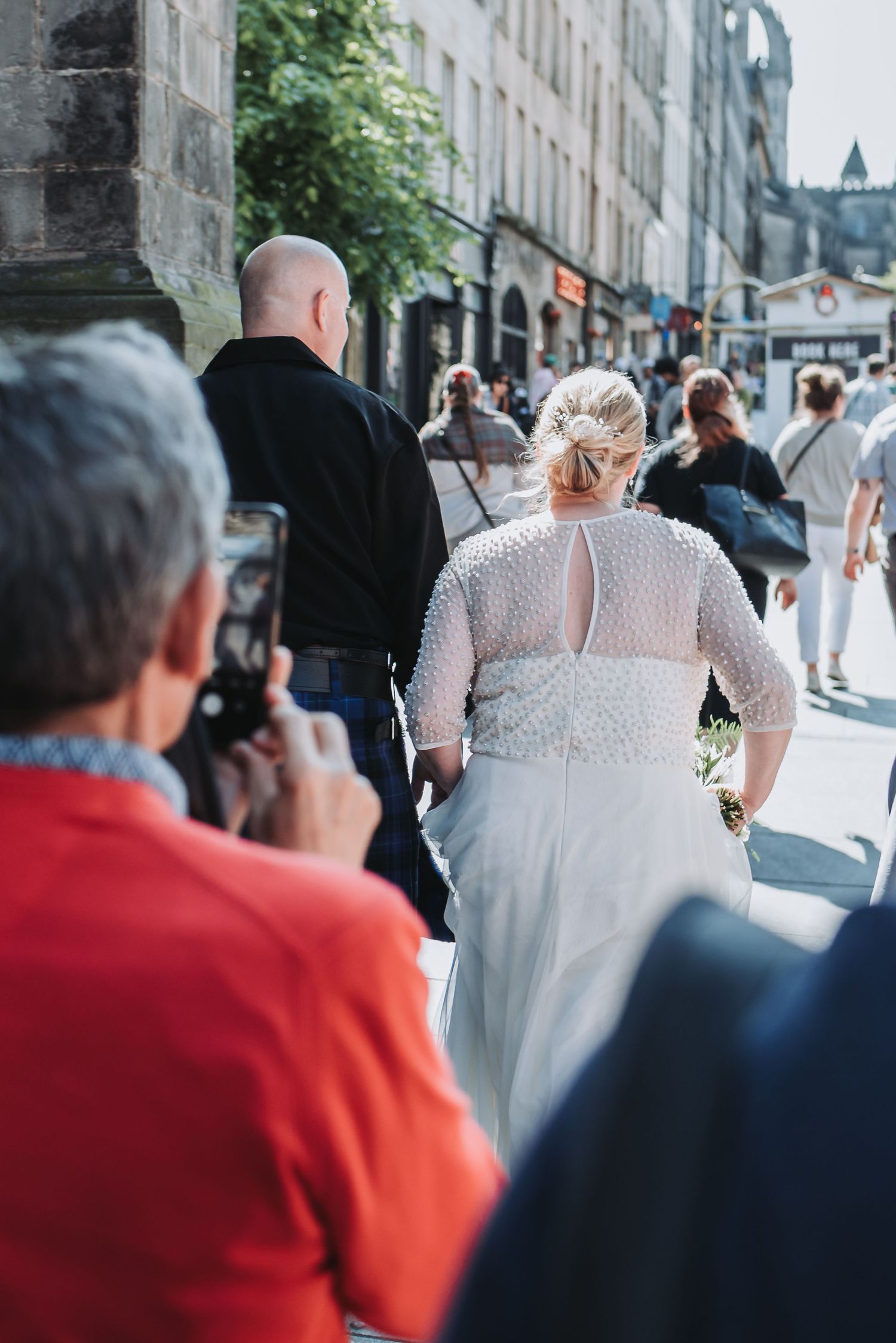 Photographer capturing bride in detailed lace dress walking through city street.