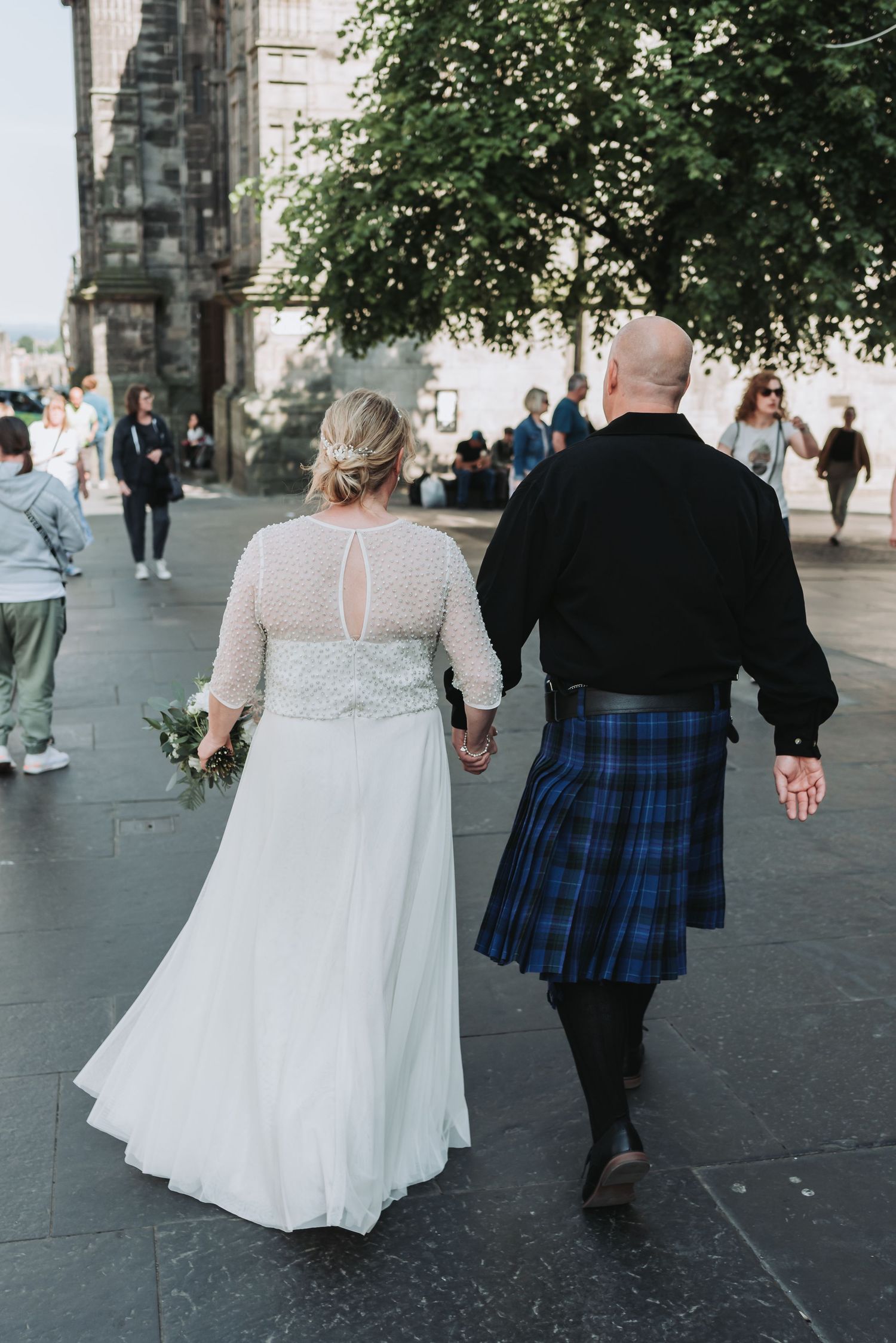 A couple in wedding attire walking hand in hand down a historic street, the groom wearing a traditional Scottish kilt.