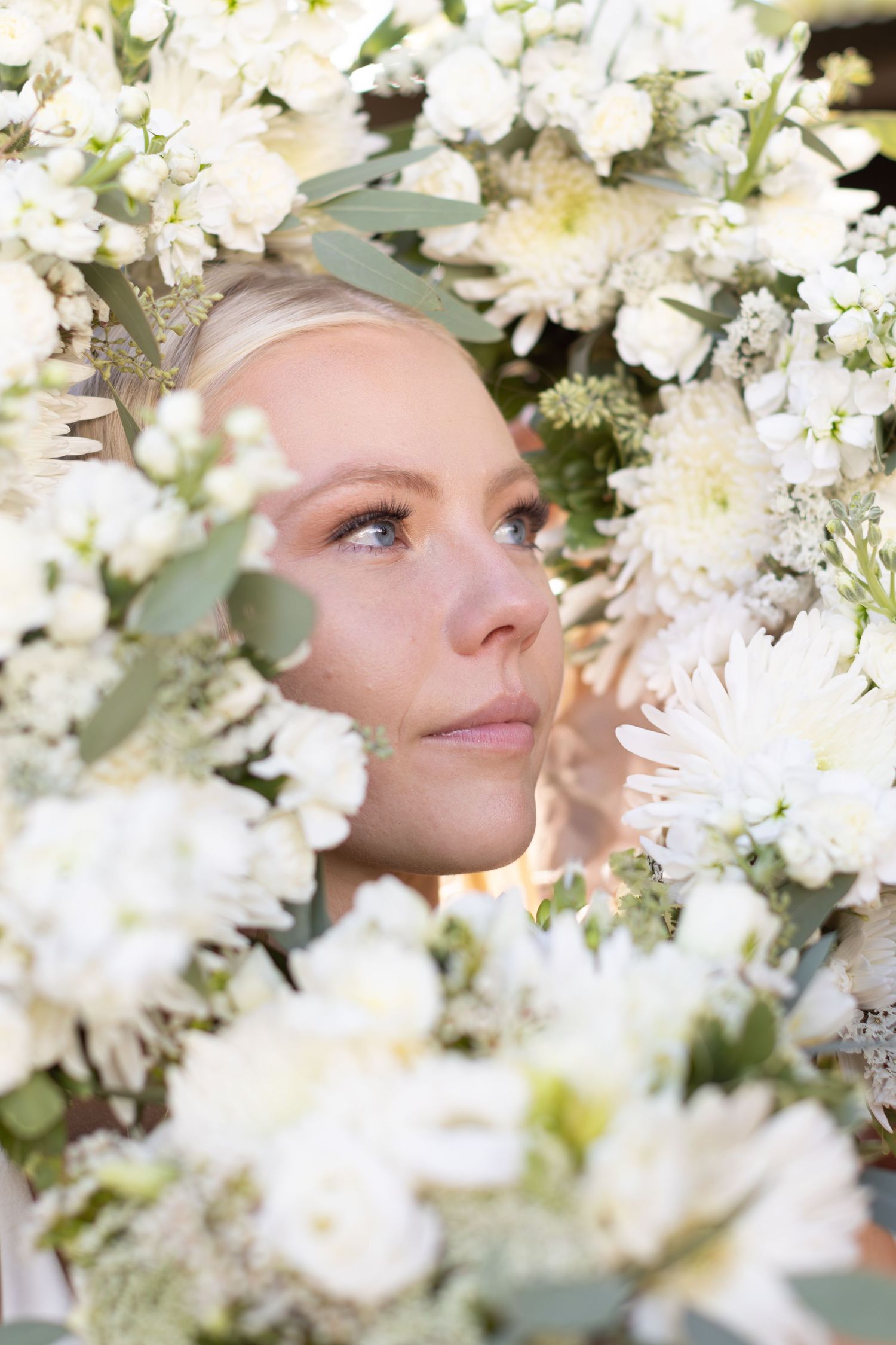 Artistic portrait framed by white flowers and eucalyptus leaves in soft lighting.