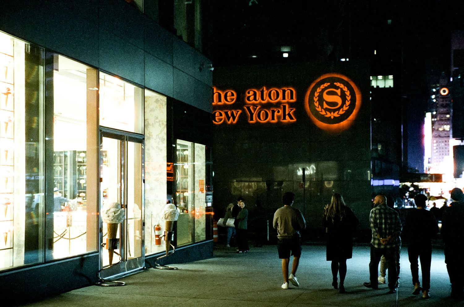 Neon sign reading 'No More No War' glows above a dark city street at night with illuminated storefronts.