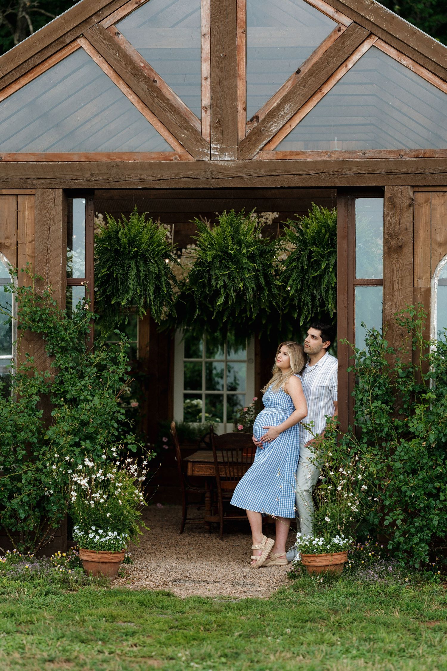 Couple poses tenderly in front of a rustic wooden greenhouse surrounded by lush greenery and flowers in Somerset, Nova Scotia.
