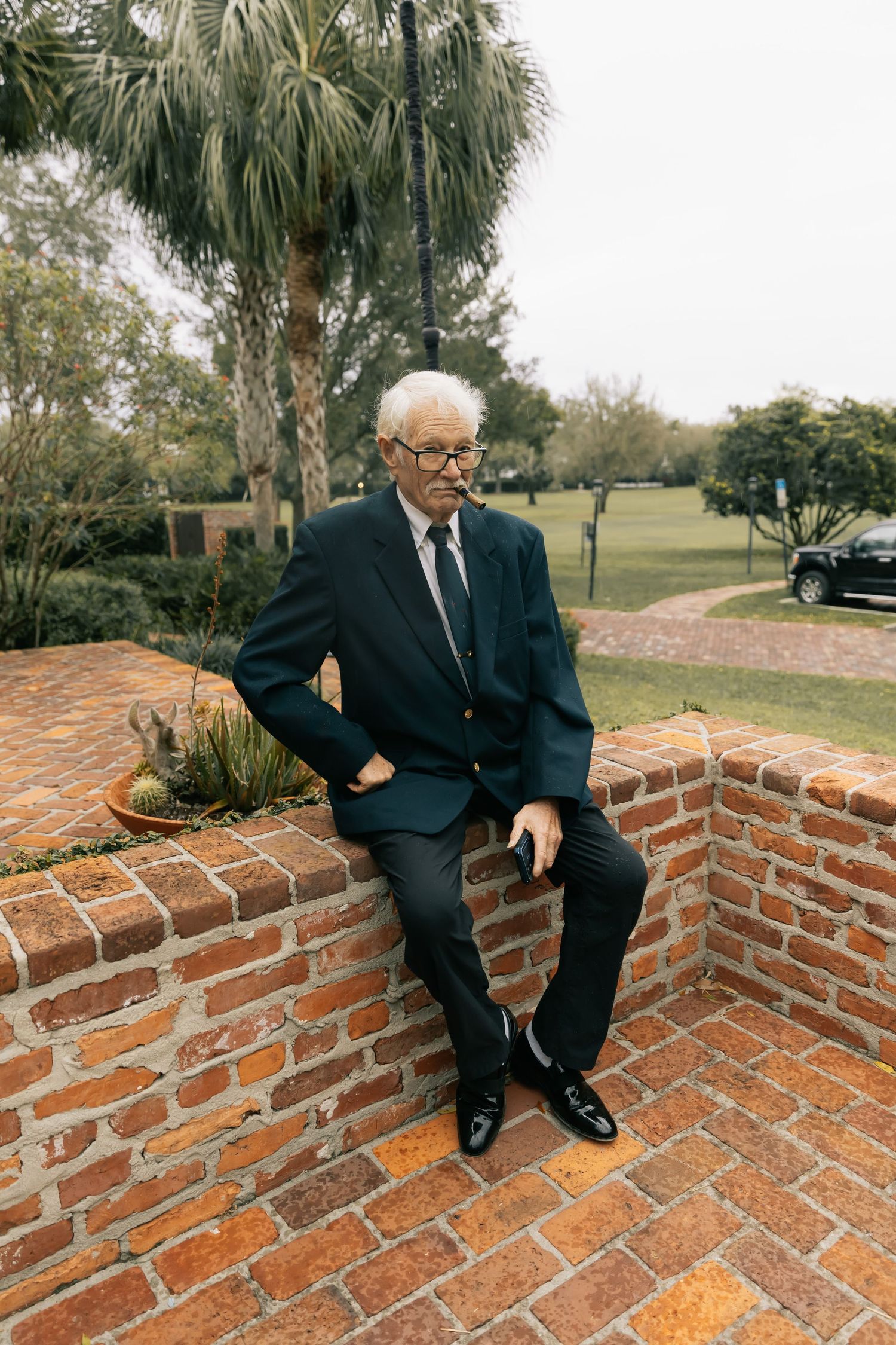 Elderly man in a navy suit, tie and glasses sits on a brick ledge in a sunny garden with palm trees behind him.