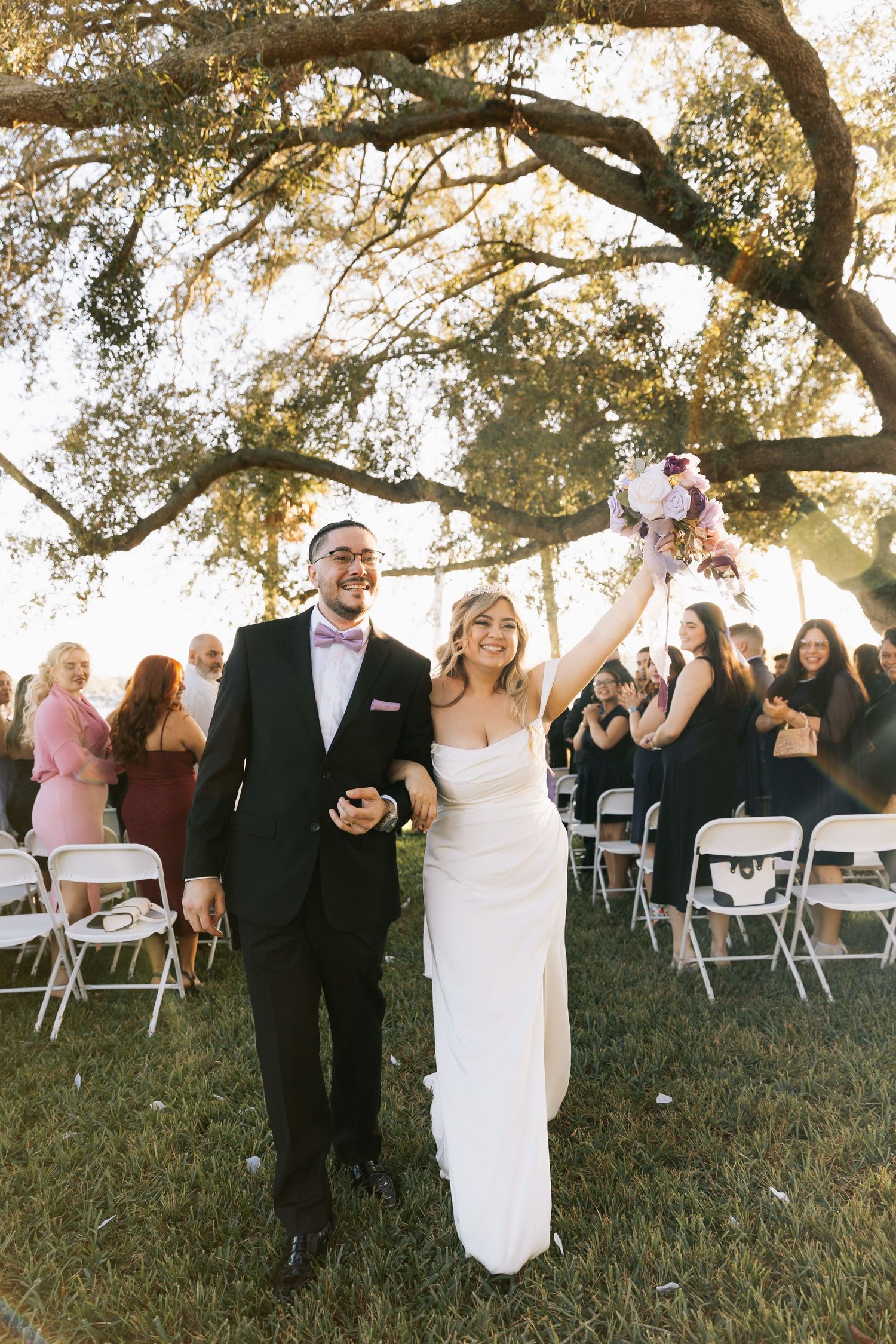 Newlyweds walk down the grass aisle past seated guests under an oak tree, the bride raising a pink and purple bouquet.