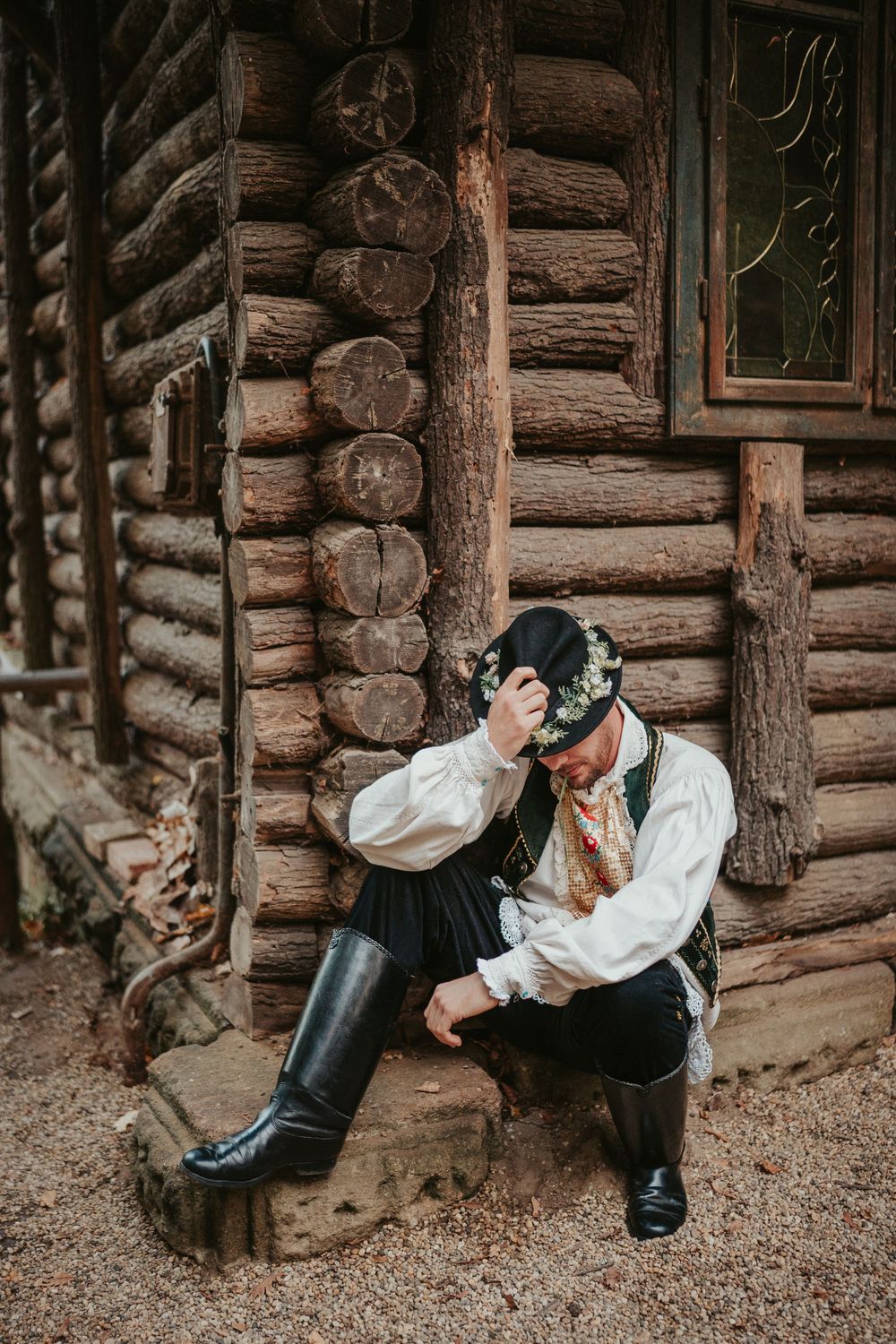 Traditional folk costume worn by person sitting against rustic log cabin wall.