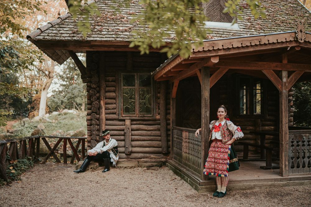 Folk-costumed figures posed by rustic wooden cabin in forest setting.