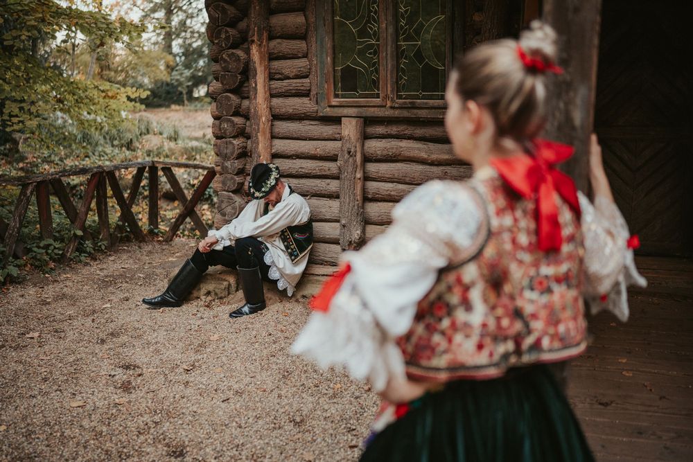 Traditional folk costumes photographed outside rustic log cabin in natural setting.