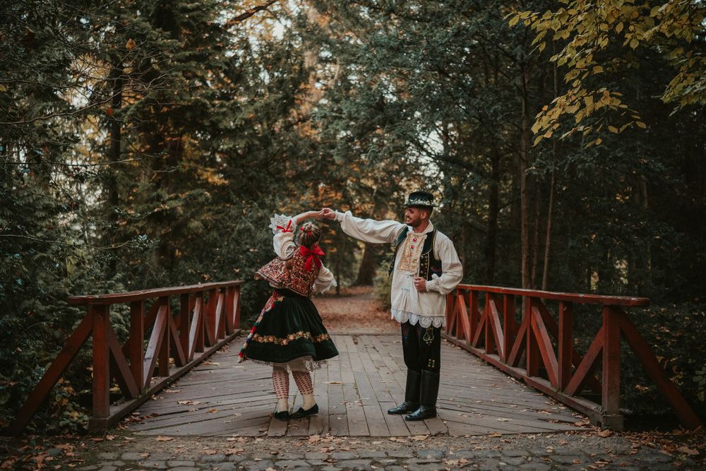 Couple in traditional folk costumes standing on wooden bridge in forest setting.
