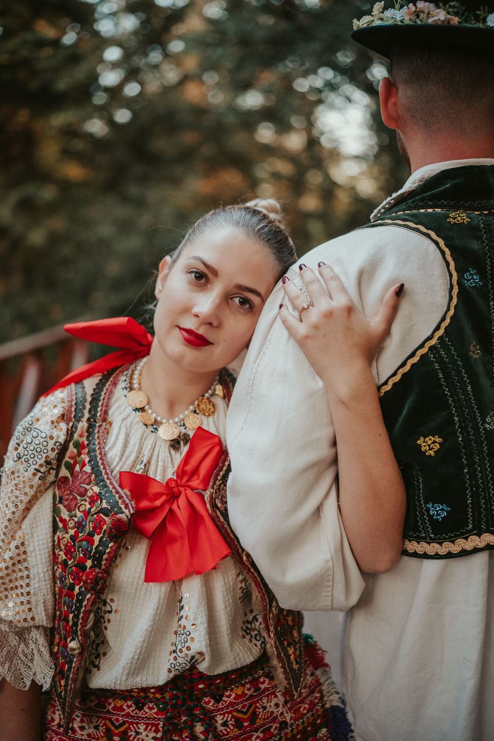 A couple in traditional Eastern European folk costumes embracing outdoors with vibrant red accents and embroidered details.