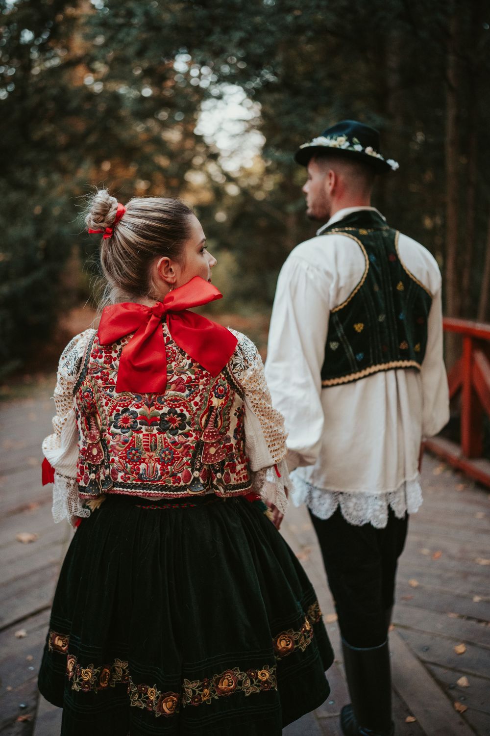 A couple in ornate folk costumes walking on a wooden bridge, with detailed embroidery and traditional accessories.