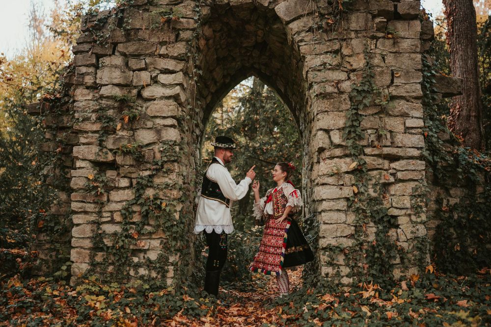 A couple in traditional folk dress posing beneath a rustic stone archway surrounded by autumn foliage.