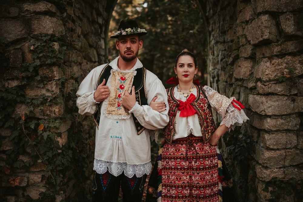 A couple in traditional Eastern European folk attire standing against a stone wall backdrop.