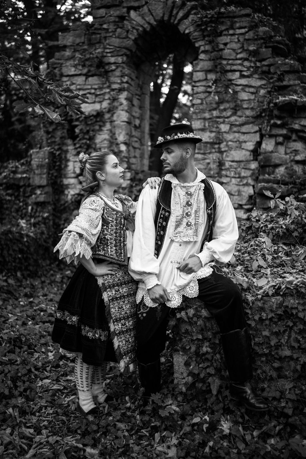 A black and white portrait of a couple in traditional folk costumes posing by a stone archway.