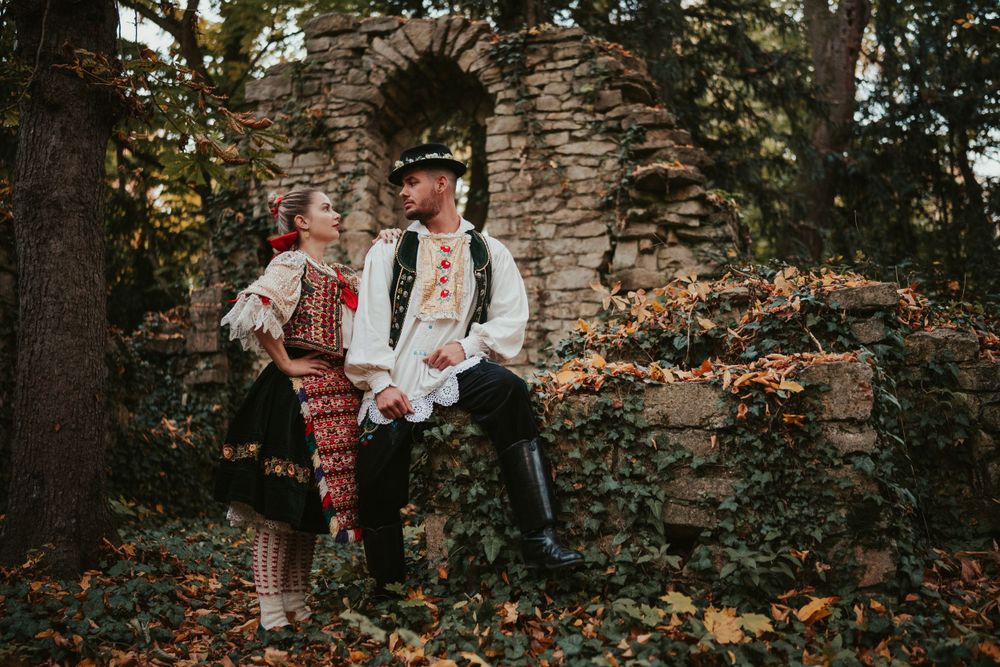 A couple in folk costumes sitting on a stone wall surrounded by autumn trees and fallen leaves.