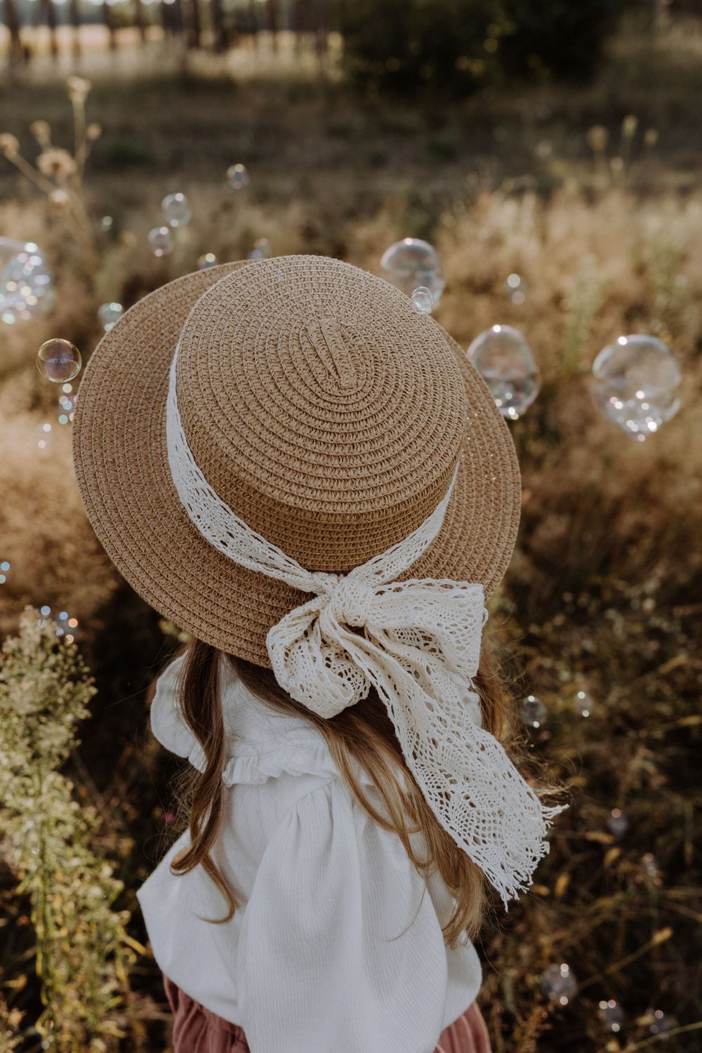 Straw sunhat with lace ribbon captured from behind with floating soap bubbles in golden light.