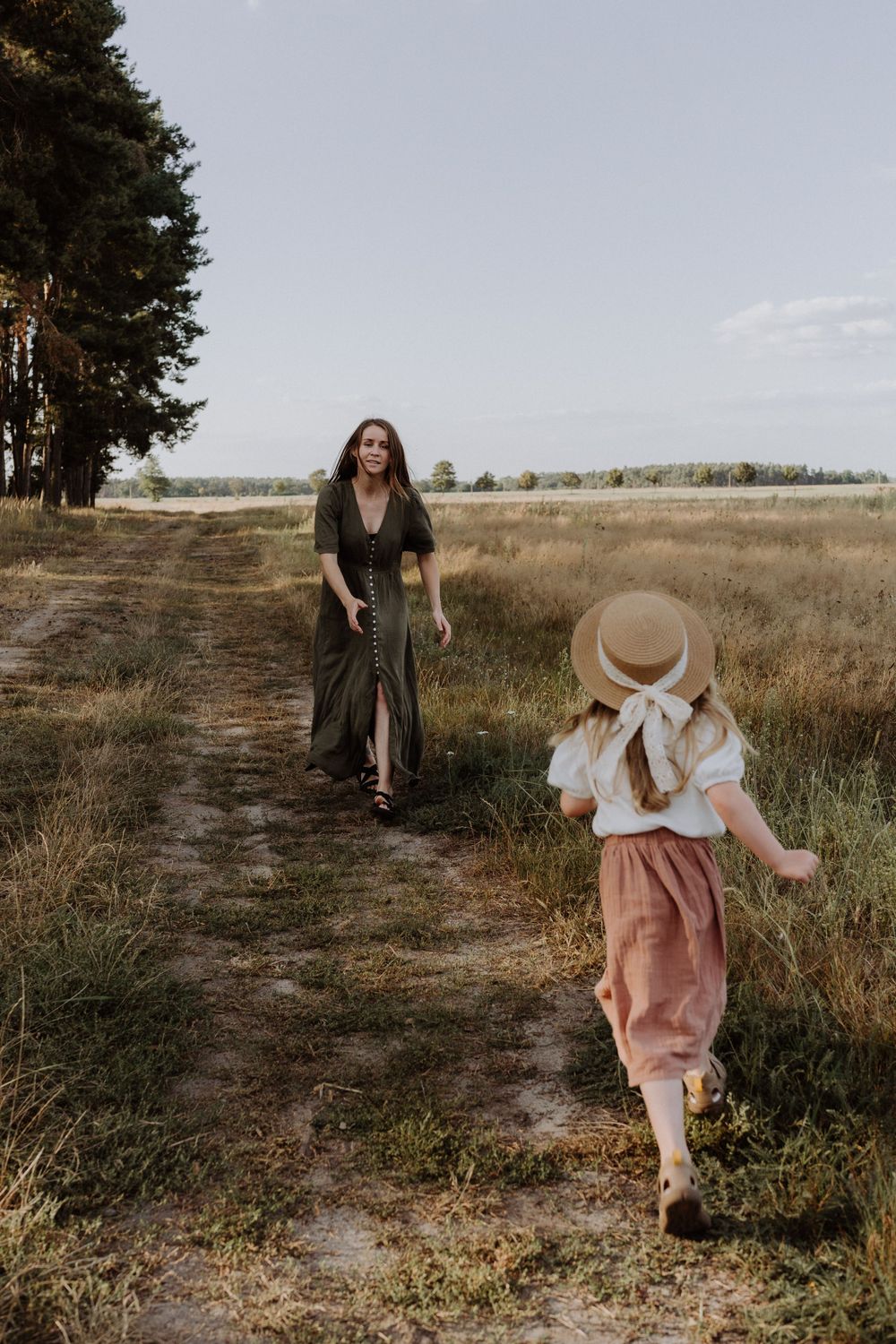 Two figures walking on a dirt path through a meadow at sunset, one wearing a straw hat and pink pants.