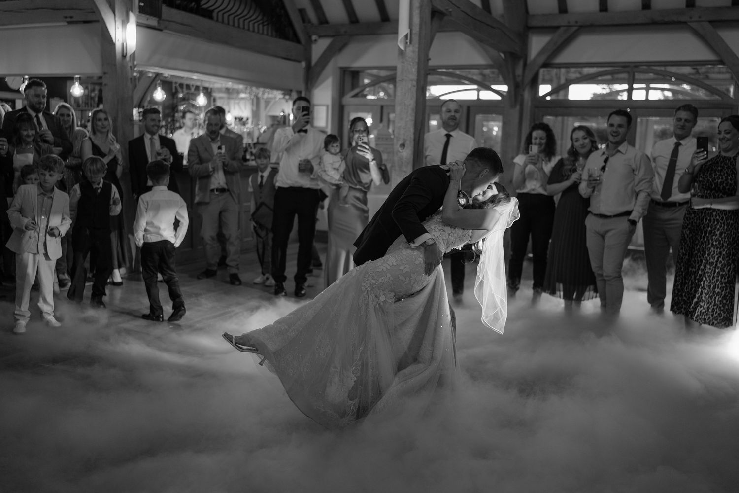 Bride and groom share romantic first dance dip surrounded by dry ice fog and wedding guests in elegant venue.