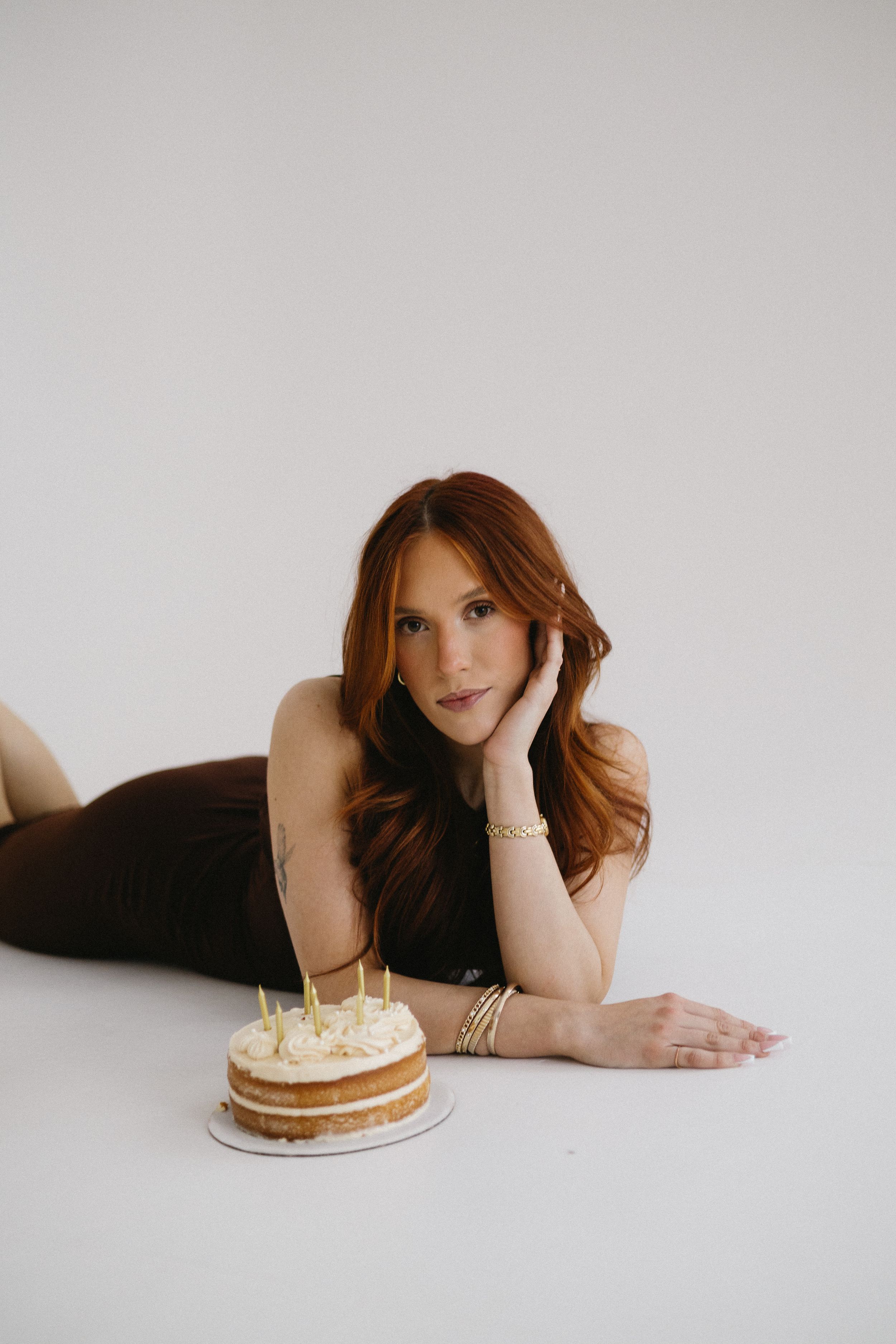 A person wearing a brown dress and laying next to a birthday cake for a birthday photography session in front of a white studio backdrop.
